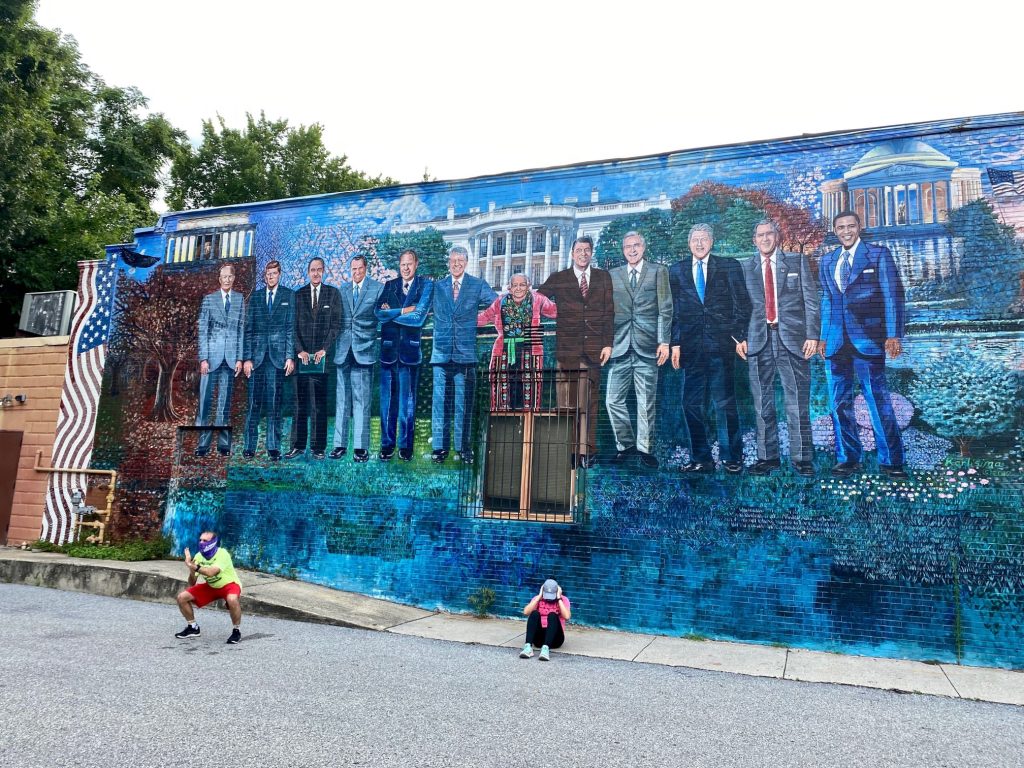 Alex and Estella workout in front of a mural with a woman standing in the middle with a pink cardigan (Mama Ayesha) and on either side of her are 11 US Presidents. 