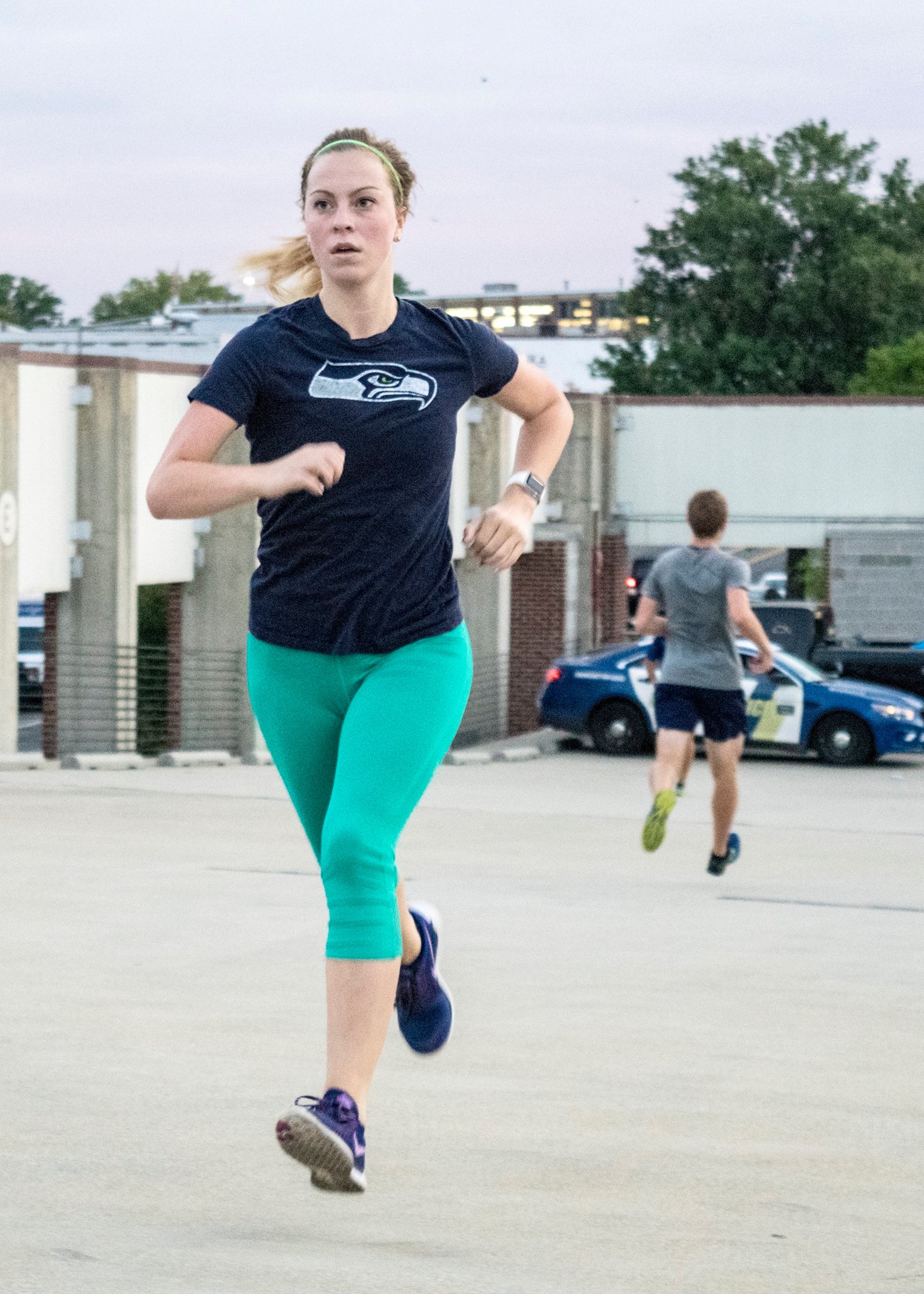 Emma is photographed in mid running stride. She is wearing purple Nike running shoes, bright teal workout pants, and a Seattle Seahawks t-shirt.