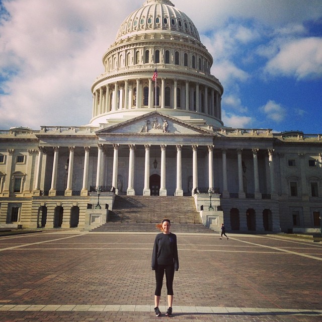 Emma stands in front of the Capitol Building in Washington, DC. She is wearing dark workout clothes and is smiling. It is a clear day and there is a bright blue sky behind the Capitol.