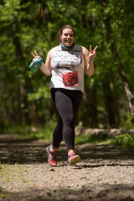 Ana running in a white teeshirt and black legging on a trail path while smiling and waving at the camera