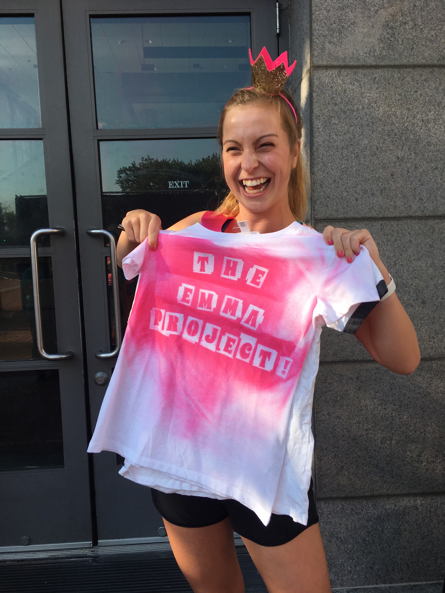 Emma is laughing and smiling while wearing a crown on her head and holding a white t-shirt with spray painted pink letters that reads, "The Emma Project."
