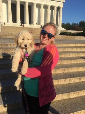 Maneula with blonde hair pulled into a pony tail holding her dog, Violet, in front of the Lincoln Memorial. Violet is a small puppy, and Manuela is wearing a pink and orang jacket with a teal tagged NP shirt. She is wearing black and blue sunglasses.
